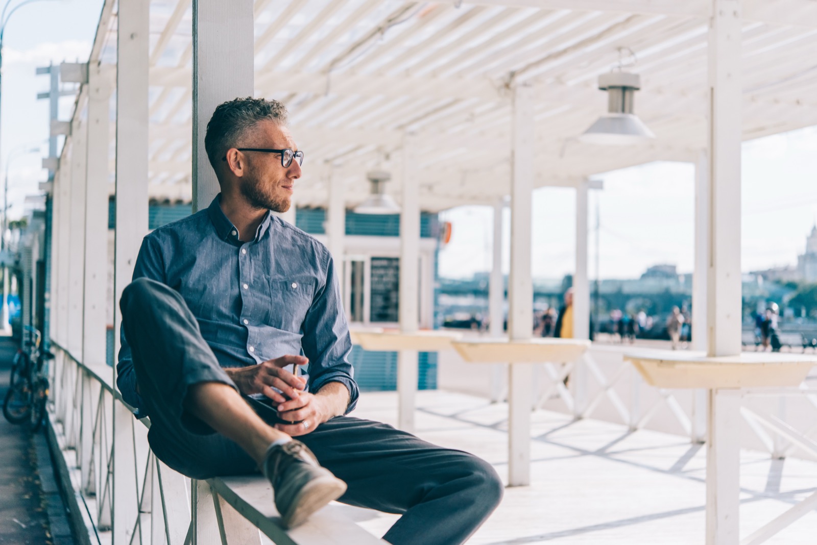A man in his fifties with graying hair and glasses sits on the railing of a sunlit terrace, looking out toward the water.